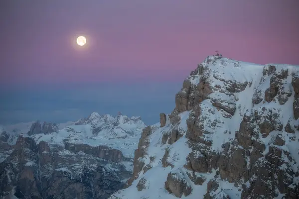 Moonrise above snowy Dolomites with hikers near the summit cross, reflecting silence, travel and mountain experiences